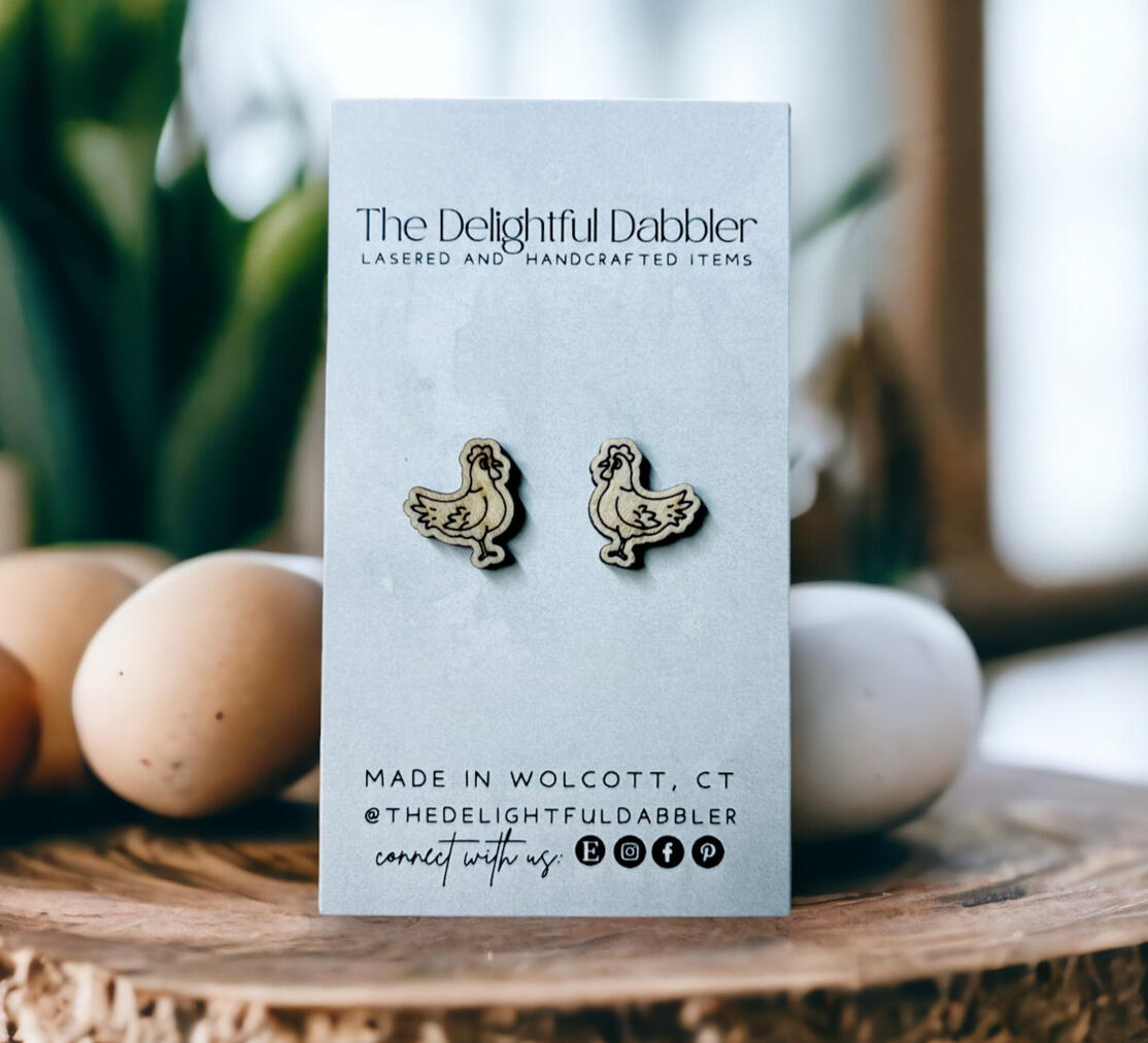 Laser cut chicken stud earrings handmade by The Delightful Dabbler, photographed with eggs on a rustic wood surface.