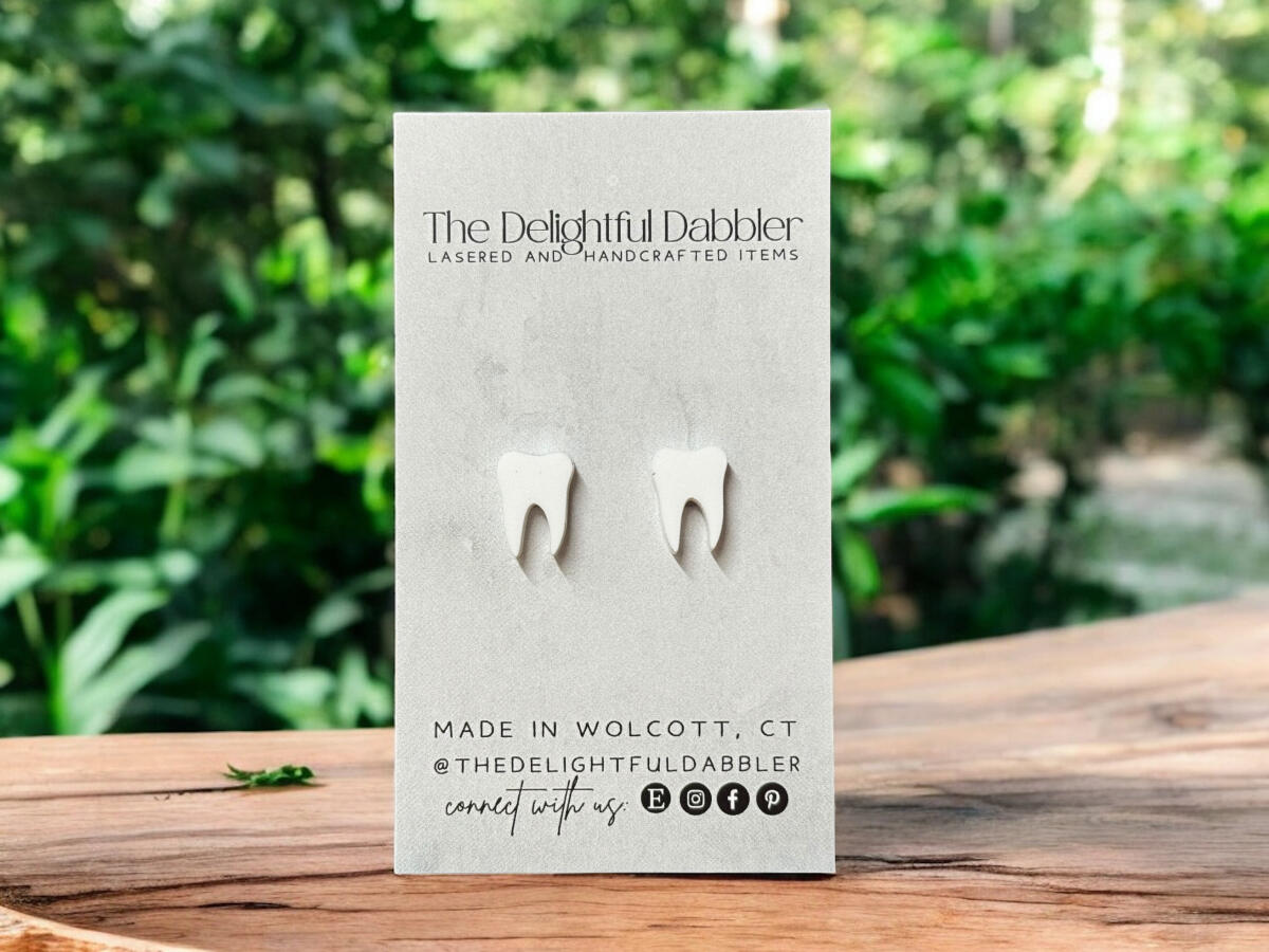 Pair of handmade matte white tooth stud earrings photographed on a branded card with a natural outdoor background.