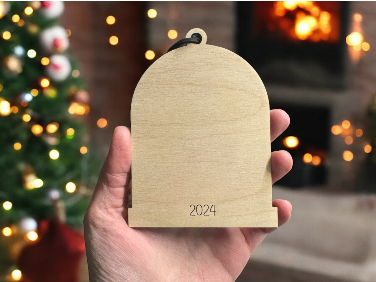 back of wood arch ornament with year engraved, being held in hand with christmas tree lights in the background